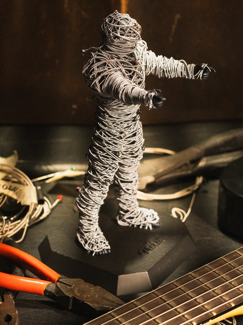 A String Thing figure fully wrapped in guitar strings, standing on a workbench beside pliers, a guitar neck, and loose coils of strings